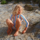 A child sits on rocks wearing a UV Protective Rash Vest Multi Stripe, looking off to the side in a thoughtful pose.